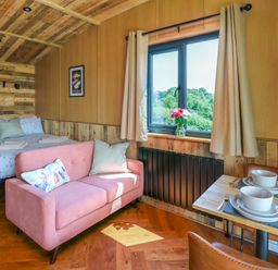 A small room with a bed a pink sofa a window with flowers and a dining table set for two at Hen's Hideaway in Rhaedr Gwy near Rhayader