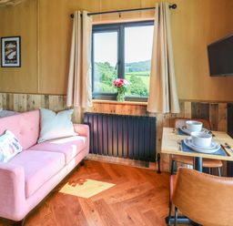 A small room with a pink sofa a window with curtains and flowers a wall-mounted television and a dining table with two chairs set for two at Hen's Hideaway in Rhaedr Gwy near Rhayader