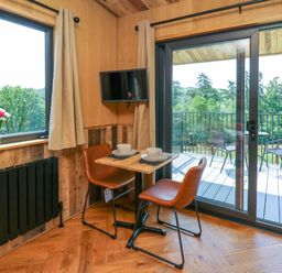 A dining area with a small table set for two and two brown chairs next to a window with flowers and a sliding door leading to a balcony with outdoor furniture at Hens Hideaway in Rhaedr Gwy near Rhayader