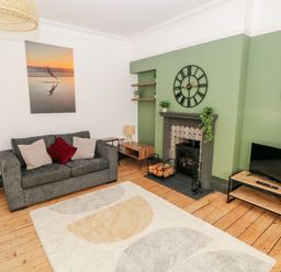 A living room with a grey sofa and cushions a fireplace with a clock above a TV on a wooden stand and a rug on a wooden floor at 36A Mary Street in Porthcawl