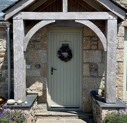A front door with a wooden porch and wreath surrounded by stone walls and plants at The Riblows Cape Cornwall Bollowal near St Just