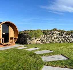 A circular wooden structure with a glass door on a stone patio in a grassy yard with a stone wall at The Riblows Cape Cornwall