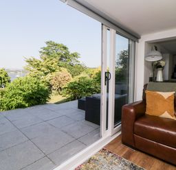 A living room with a brown leather sofa next to large sliding glass doors opening to a patio and garden at Riverview @ 1 Brunel View in Saltash