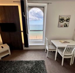 A room with a window overlooking the beach and sea a wall-mounted TV a white dining table with three chairs and a floor lamp at Apartment 9 Beaconsfield House Bridlington