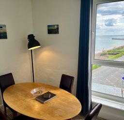 A dining room with a wooden table and four chairs near a window overlooking a coastal road at 7 Beach View @ Beaconsfield House in Bridlington
