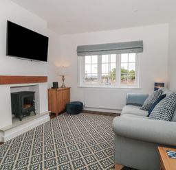 A living room with a wall-mounted television a small wood stove a window with a view a sofa and a patterned rug at Quay Cottage - Sea View in Poole