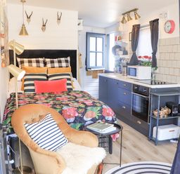 A small room with a bed with multiple pillows a floral quilt a kitchen counter with blue cabinets a microwave oven a chair with a striped cushion a floor lamp and wall decorations at Creed Cabin in Stornoway