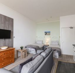 A room with two single beds a sofa a wooden chest of drawers a wall-mounted TV and a kitchenette at 4 Beach View Beaconsfield House in Bridlington