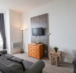 A living room with a gray sofa a wooden chest of drawers a small table with a plant a floor lamp and a wall-mounted TV at 4 Beach View Beaconsfield House Bridlington