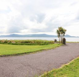 A paved driveway leading to a gate with palm trees beside a grassy area and a body of water with hills in the background at Meeligolleen in Coill Tra