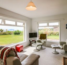 A living room with large windows showing a garden and hills with white leather chairs and a stone fireplace at Meeligolleen in Coill Tra