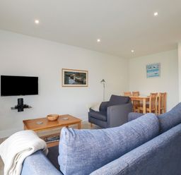 A living room with blue couches a wooden coffee table a wall-mounted television and a dining table with chairs at Freefolk Cottage in Polzeath