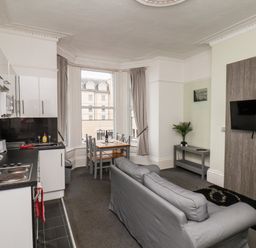 An open plan kitchen and living room with a gray sofa a wall mounted television a dining table with chairs and large windows at Promenade in Bridlington