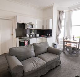 A living area with a gray sofa, a kitchen with white cabinets and appliances, and a dining table with four chairs by large windows at Promenade Bridlington