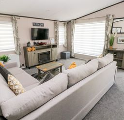A living room with two gray sofas a wooden TV stand with a fireplace a wall-mounted TV and large windows with curtains at Beechwood Lodge in Hasguard Cross near Broad Haven