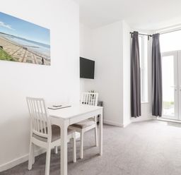 A dining area with a white table and two chairs next to a wall-mounted TV and large windows with curtains at 3 Seaview @ Bridlington Bay in Bridlington