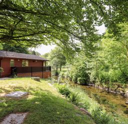 A wooden lodge next to a stream surrounded by grass and trees at Nutkins Lodge Beckside 5 Limefitt Park near Windermere