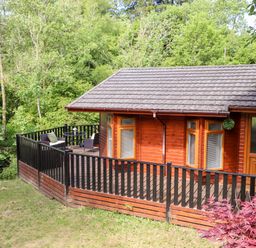 A wooden cabin with a black and brown railing and outdoor seating surrounded by trees at Nutkins Lodge Beckside 5 Limefitt Park near Windermere