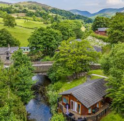 A wooden cabin near a river with a stone bridge and green hills in the background at Nutkins Lodge Beckside 5 Limefitt Park near Windermere
