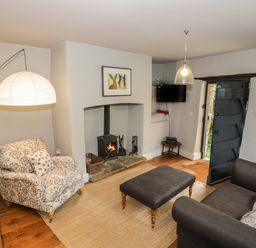A living room with a wood stove fireplace a patterned armchair a dark sofa a large ottoman and an arched floor lamp at The Old Brewhouse in Chipping Campden