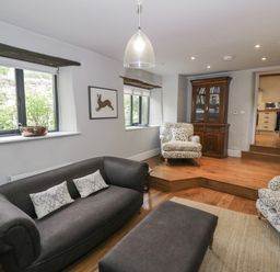 A living room with a dark grey sofa, two patterned armchairs, wooden cabinet, and windows at The Old Brewhouse in Chipping Campden