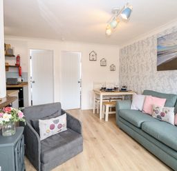 A living area with a grey armchair and a teal sofa with cushions a wooden dining table set against a wall with leaf pattern wallpaper at 72 in Hemsby