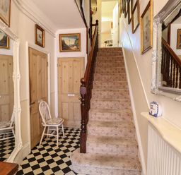 A hallway with a carpeted staircase a white chair checkered floor wooden doors and framed pictures at The Studio in Sheringham