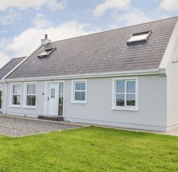 A single story house with grey walls, a dark grey roof, white framed windows and a white door surrounded by grass at The Haven in Falcarragh County Donegal