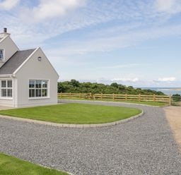 A house with white walls and gray roof by a gravel driveway and green lawn near a wooden fence with water and bushes in the background at The Haven in Falcarragh County Donegal