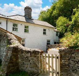 A white stone house with a slate roof behind a stone wall and wooden gate with greenery around at Fron Dderw Bala