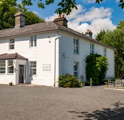 The exterior of a white two-story house with multiple windows and a gravel driveway outdoor seating area with tables and chairs surrounded by trees and greenery at Fron Dderw in Bala