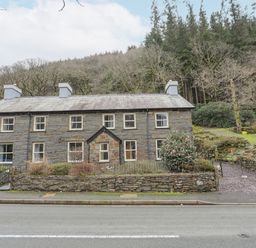 A stone cottage with multiple chimneys and windows by a road near a wooded hill at Glyn Awel in Nant Gwynant near Beddgelert
