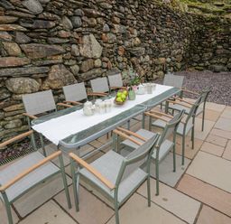 Outdoor patio area with a glass table and eight chairs against a stone wall at Glyn Awel in Nant Gwynant near Beddgelert