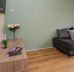 A corner of a living room with a wooden cabinet holding a vase of white flowers a bottle of champagne two glasses and magazines next to a dark gray sofa at Bwythyn Yr Wyddfa in Llanberis
