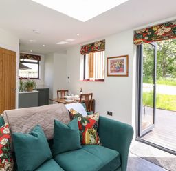 A living area with green sofa and floral cushions next to a dining table and open door to garden at Brow Wood Cabin in Ullswater