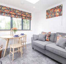 A living room with a grey sofa, wooden chairs, a round wooden table with dishes and flowers and floral curtains at Bluebell Cabin in Ullswater
