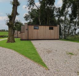 A wooden cabin with gravel driveway and green grass surrounded by trees at Bluebell Cabin in Ullswater