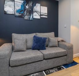 A living room with a gray sofa with cushions a side table with a table lamp a floor lamp and abstract paintings on a dark wall at The Dollhouse in Cayton Bay