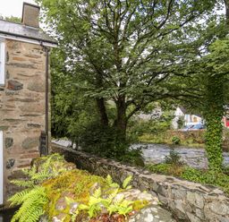 A stone house next to a river with trees and a stone wall with moss and ferns at Ty Nain in Beddgelert