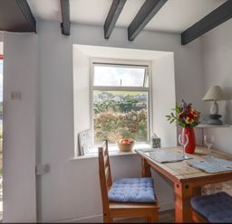 A dining area with a wooden table and chairs with blue cushions a flower vase and window showing outdoor view at Chy Rani in Portmellon near Mevagissey