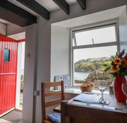 A kitchen dining area with a red door open to the outside and a window with a view of the coastline at Chy Rani in Portmellon near Mevagissey