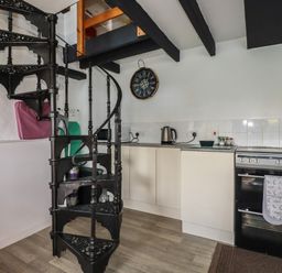 A kitchen with a black spiral staircase a black oven cream cabinets a kettle on the countertop and a wall clock at Chy Rani in Portmellon near Mevagissey