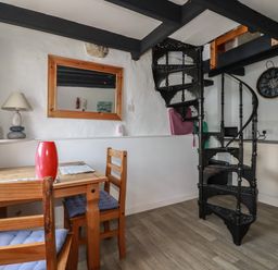 A dining area with a wooden table and chairs a red vase a table lamp a mirror on the wall a black spiral staircase and a kitchen counter with a kettle at Chy Rani in Portmellon near Mevagissey