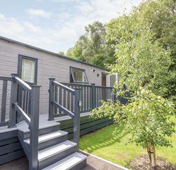 A raised wooden deck with stairs leading to a grey mobile home surrounded by green grass and trees at 7 Wild Haven in Landford