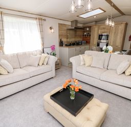 A living room with two beige sofas a beige ottoman with flowers and a kitchen in the background at 7 Wild Haven in Landford