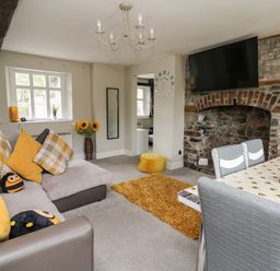 A living room with a sofa and yellow pillows a dining table with chairs a wall clock and a television at Yewtree Cottage in Defynog near Sennybridge