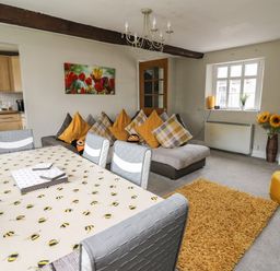 A living room with a dining table covered with a bee-patterned tablecloth chairs a sofa with yellow and plaid cushions a yellow rug and a yellow pouf at Yewtree Cottage in Defynog near Sennybridge