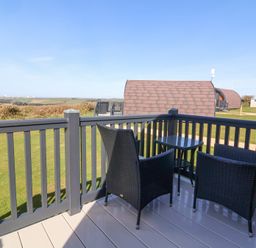 An outdoor deck with two black chairs and a small table overlooking grassy area and brown roofed accommodation at Owl's Nook in Kilkhampton