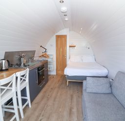 A narrow room with a bed a gray sofa a kitchenette with stove oven and sink and two white chairs at Owls Nook in Kilkhampton