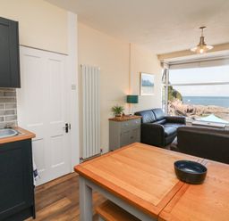 A living area with a wooden table a black bowl two black sofas a sideboard with a lamp and plant a kitchen sink and cabinets and a window showing a coastal view at At the Bay Apartment in Hele Bay Ilfracombe